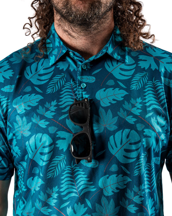 Men’s Cabana Nights golf polo by Pins & Aces featuring a blue and teal tropical leaf print, short sleeves, and athletic performance fit.