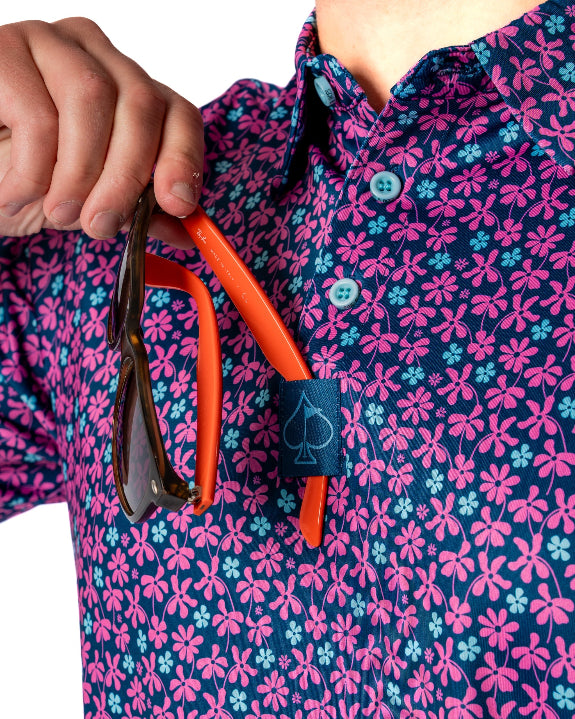 Men’s Full Bloom golf polo by Pins & Aces featuring an all-over bright pink floral pattern on a navy background with short sleeves and an athletic performance fit.