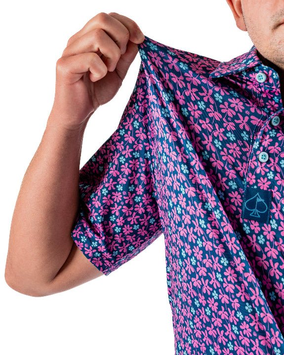 Men’s Full Bloom golf polo by Pins & Aces featuring an all-over bright pink floral pattern on a navy background with short sleeves and an athletic performance fit.