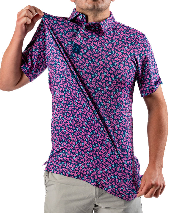 Men’s Full Bloom golf polo by Pins & Aces featuring an all-over bright pink floral pattern on a navy background with short sleeves and an athletic performance fit.