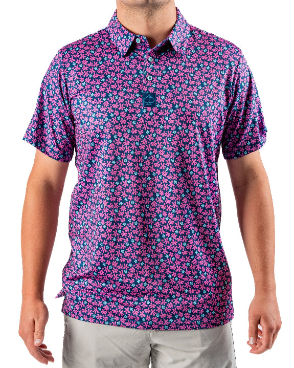 Men’s Full Bloom golf polo by Pins & Aces featuring an all-over bright pink floral pattern on a navy background with short sleeves and an athletic performance fit.