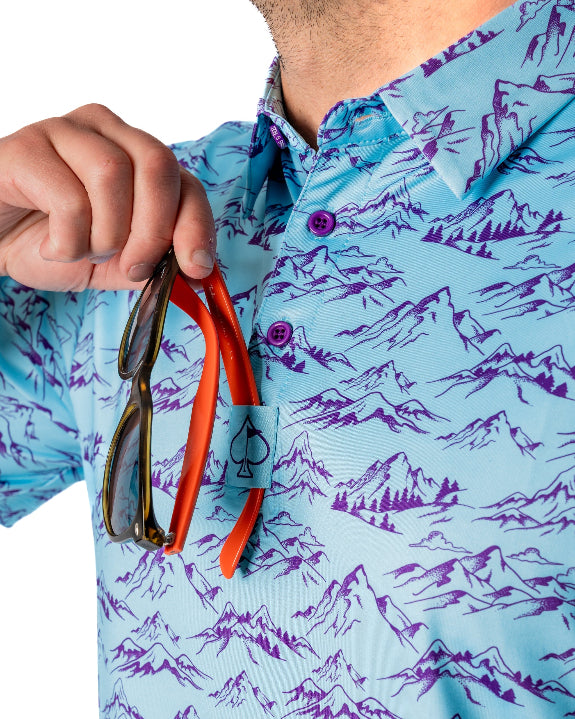 Men’s Purple Majesty golf polo by Pins & Aces featuring an aqua blue base with an all-over purple illustrated mountain print, short sleeves, and athletic performance fit.