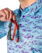 Men’s Purple Majesty golf polo by Pins & Aces featuring an aqua blue base with an all-over purple illustrated mountain print, short sleeves, and athletic performance fit.