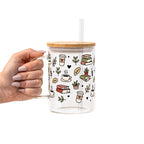 Clear glass mug with bamboo lid and straw featuring a wraparound book lover design with stacked books, coffee cups, potted plants, leafy branches and small black hearts.
