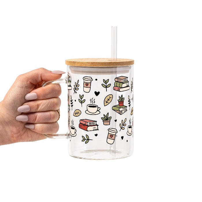 Clear glass mug with bamboo lid and straw featuring a wraparound book lover design with stacked books, coffee cups, potted plants, leafy branches and small black hearts.