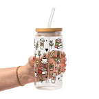 Clear glass mug with bamboo lid and straw featuring a wraparound book lover design with stacked books, coffee cups, potted plants, leafy branches and small black hearts.