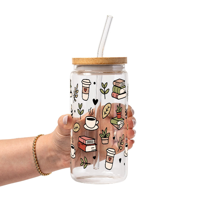 Clear glass mug with bamboo lid and straw featuring a wraparound book lover design with stacked books, coffee cups, potted plants, leafy branches and small black hearts.