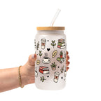 Clear glass mug with bamboo lid and straw featuring a wraparound book lover design with stacked books, coffee cups, potted plants, leafy branches and small black hearts.