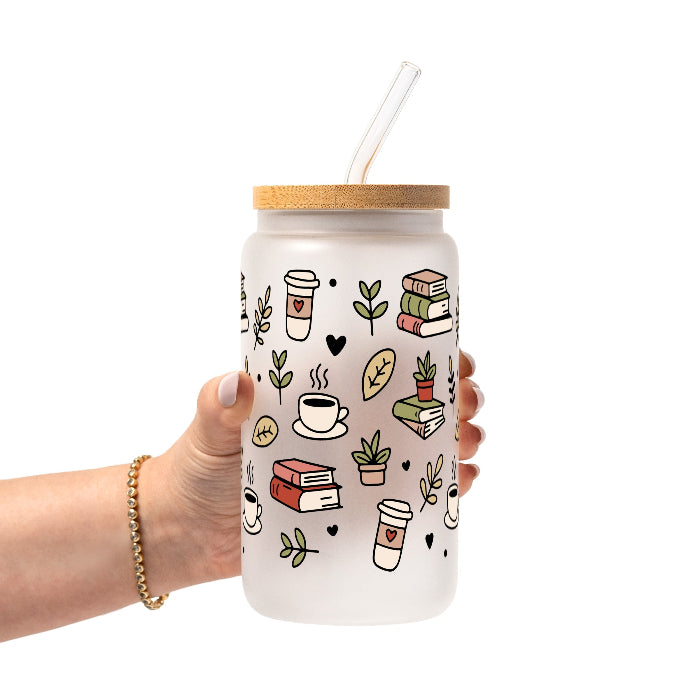 Clear glass mug with bamboo lid and straw featuring a wraparound book lover design with stacked books, coffee cups, potted plants, leafy branches and small black hearts.