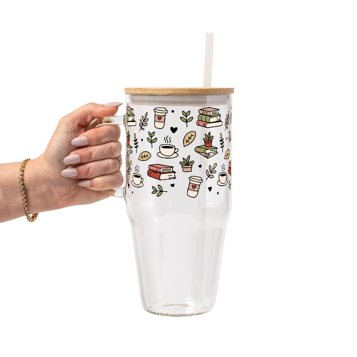 Clear glass mug with bamboo lid and straw featuring a wraparound book lover design with stacked books, coffee cups, potted plants, leafy branches and small black hearts.
