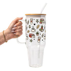 Clear glass mug with bamboo lid and straw featuring a wraparound book lover design with stacked books, coffee cups, potted plants, leafy branches and small black hearts.