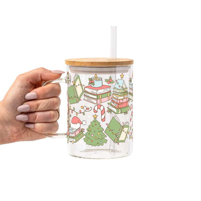 Clear glass cup with bamboo lid and straw featuring a festive wraparound design of stacked books, decorated Christmas trees, candy canes, candles, holly and colorful holiday accents.