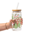 Clear glass cup with bamboo lid and straw featuring a festive wraparound design of stacked books, decorated Christmas trees, candy canes, candles, holly and colorful holiday accents.