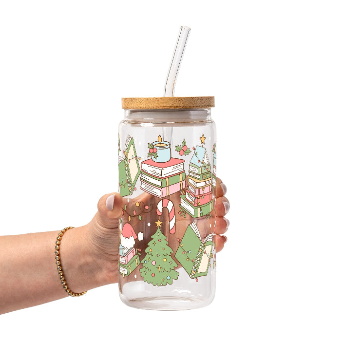 Clear glass cup with bamboo lid and straw featuring a festive wraparound design of stacked books, decorated Christmas trees, candy canes, candles, holly and colorful holiday accents.