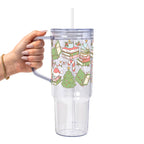 Clear glass cup with bamboo lid and straw featuring a festive wraparound design of stacked books, decorated Christmas trees, candy canes, candles, holly and colorful holiday accents.