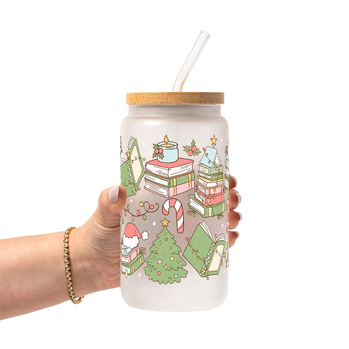 Clear glass cup with bamboo lid and straw featuring a festive wraparound design of stacked books, decorated Christmas trees, candy canes, candles, holly and colorful holiday accents.