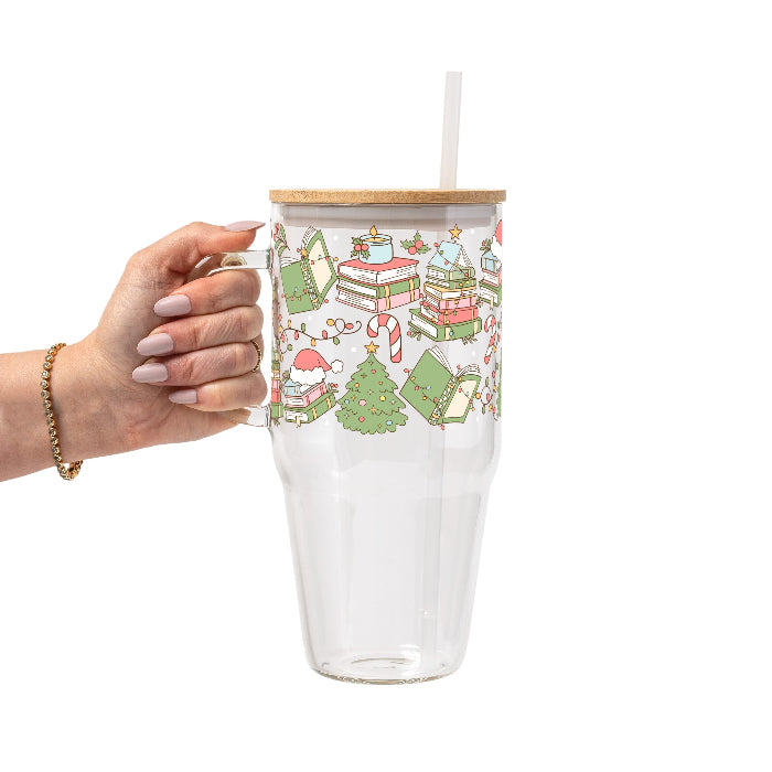 Clear glass cup with bamboo lid and straw featuring a festive wraparound design of stacked books, decorated Christmas trees, candy canes, candles, holly and colorful holiday accents.