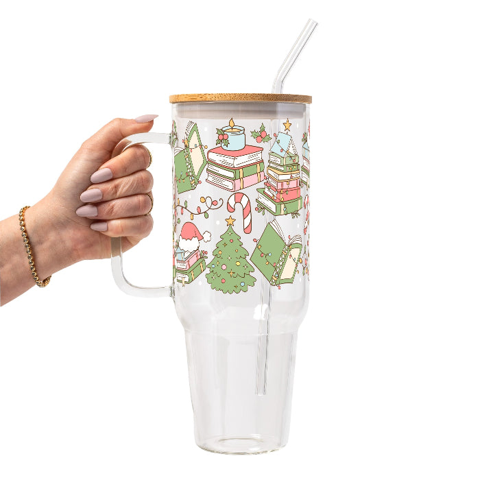 Clear glass cup with bamboo lid and straw featuring a festive wraparound design of stacked books, decorated Christmas trees, candy canes, candles, holly and colorful holiday accents.