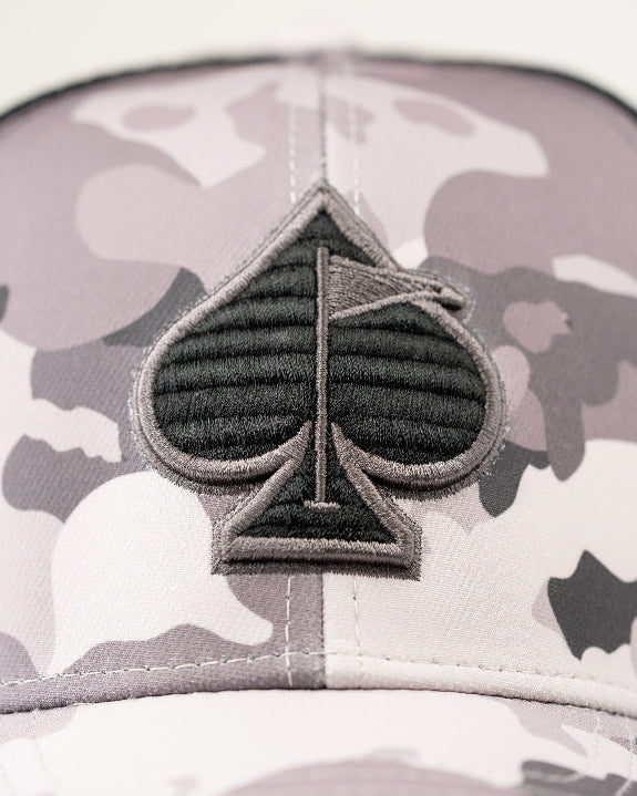 Urban Camo trucker golf hat by Pins & Aces featuring a gray, black, and white camouflage print, embroidered black spade logo, black mesh back, curved bill, and snapback design.